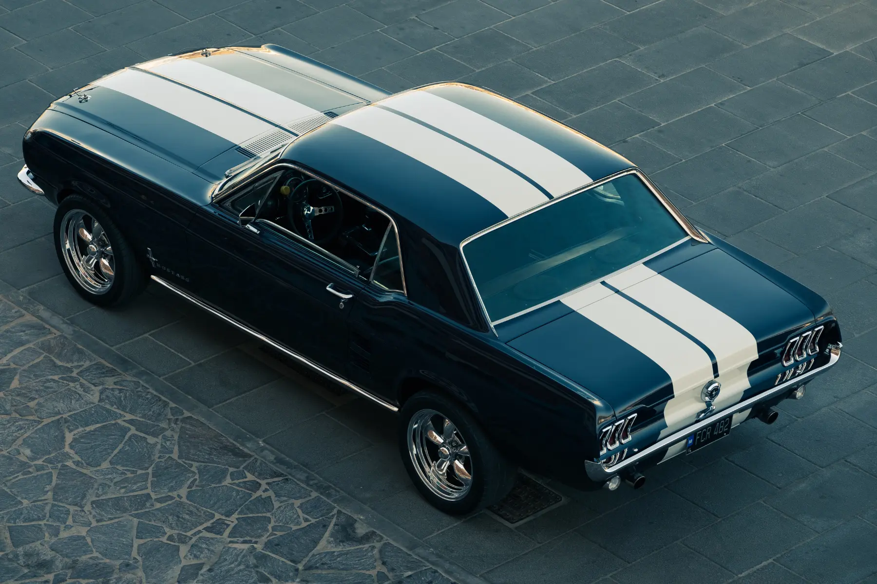 Overhead view of a classic navy-blue Ford Mustang with white racing stripes parked on a stone pavement